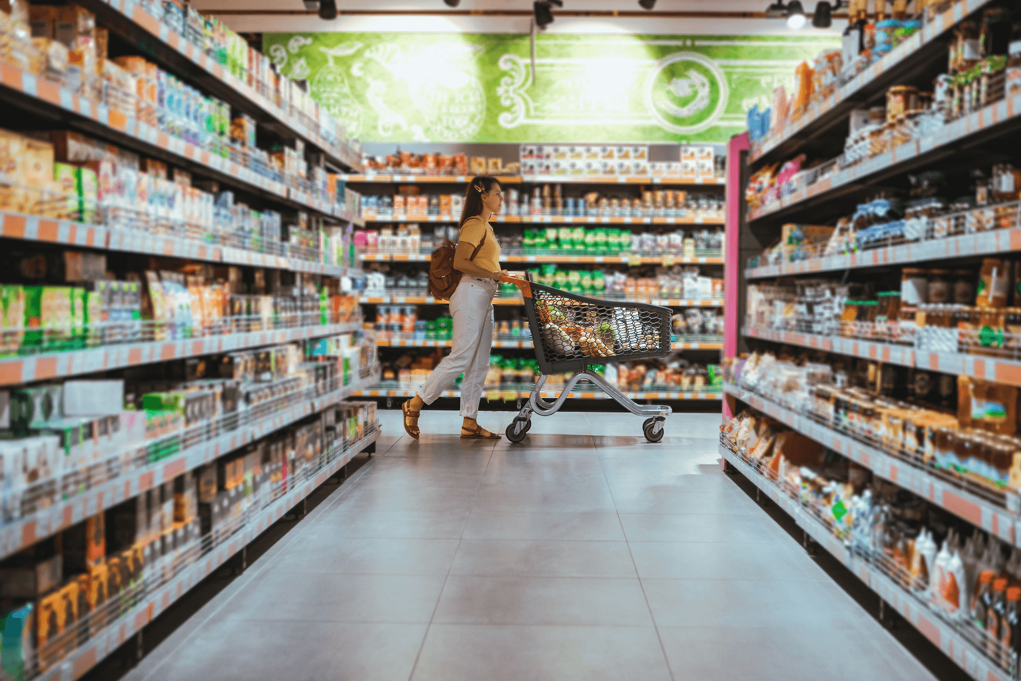 Woman pushing a shopping cart down a grocery store aisle filled with various packaged goods.