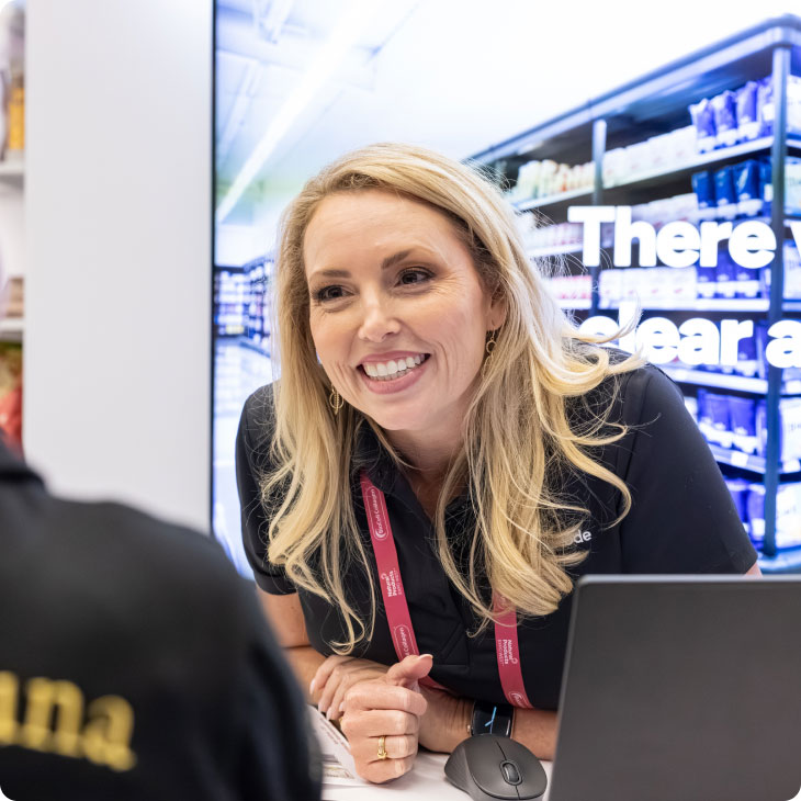 Smiling woman with blonde hair wearing a black shirt and lanyard, talking to a person across a desk.
