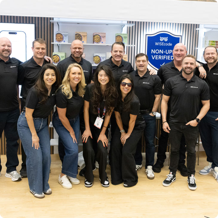 Group of nine employees smiling wearing black shirts posing at a trade show with shelves of products.