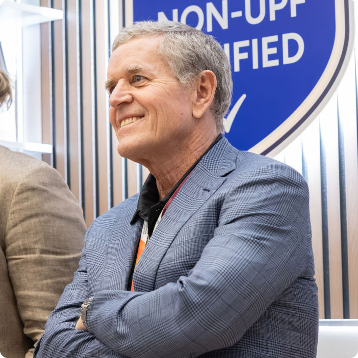 CEO smiling in a suit jacket sitting with arms crossed near a sign saying NON-UPF CERTIFIED.