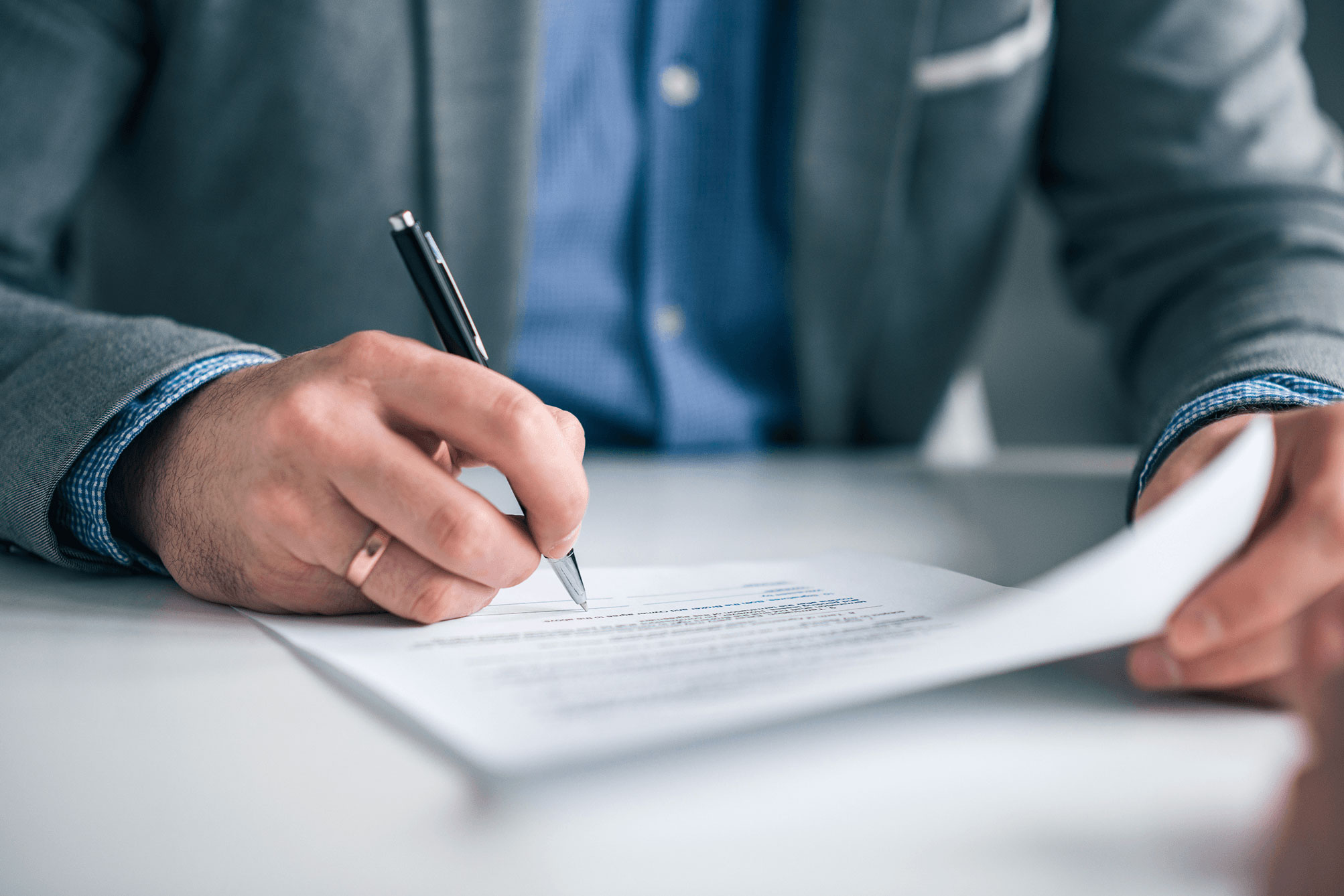 Man's hand with a wedding ring holding a pen, writing on a document on a white table.