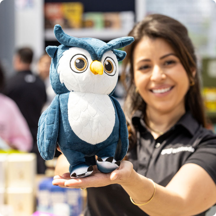Woman smiling and holding a blue and white owl plush toy with large eyes and a yellow beak.