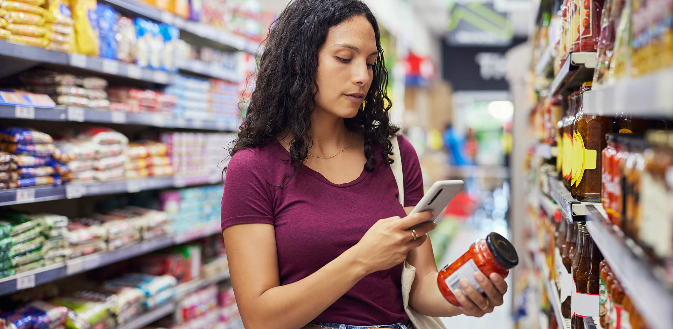Woman checking a jarred product against her phone while grocery shopping