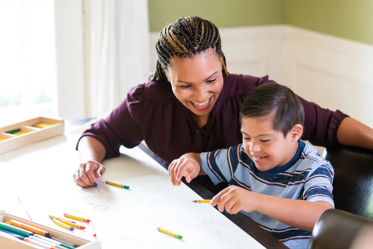 Educator smiling while drawing alongside a young student at a home table