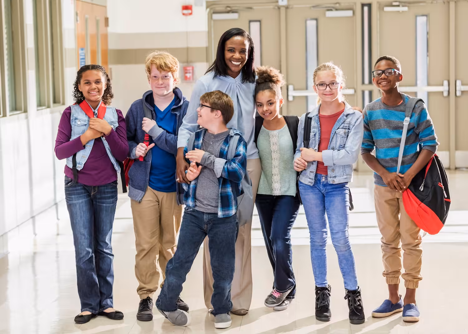 Teacher smiling with a diverse group of six elementary students with backpacks in a school hallway