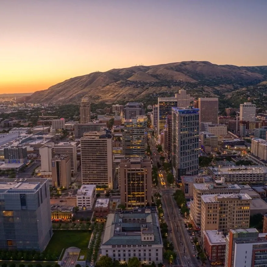 Aerial view of a cityscape at sunset with tall buildings and streets leading toward a large mountain in the background under a clear sky.