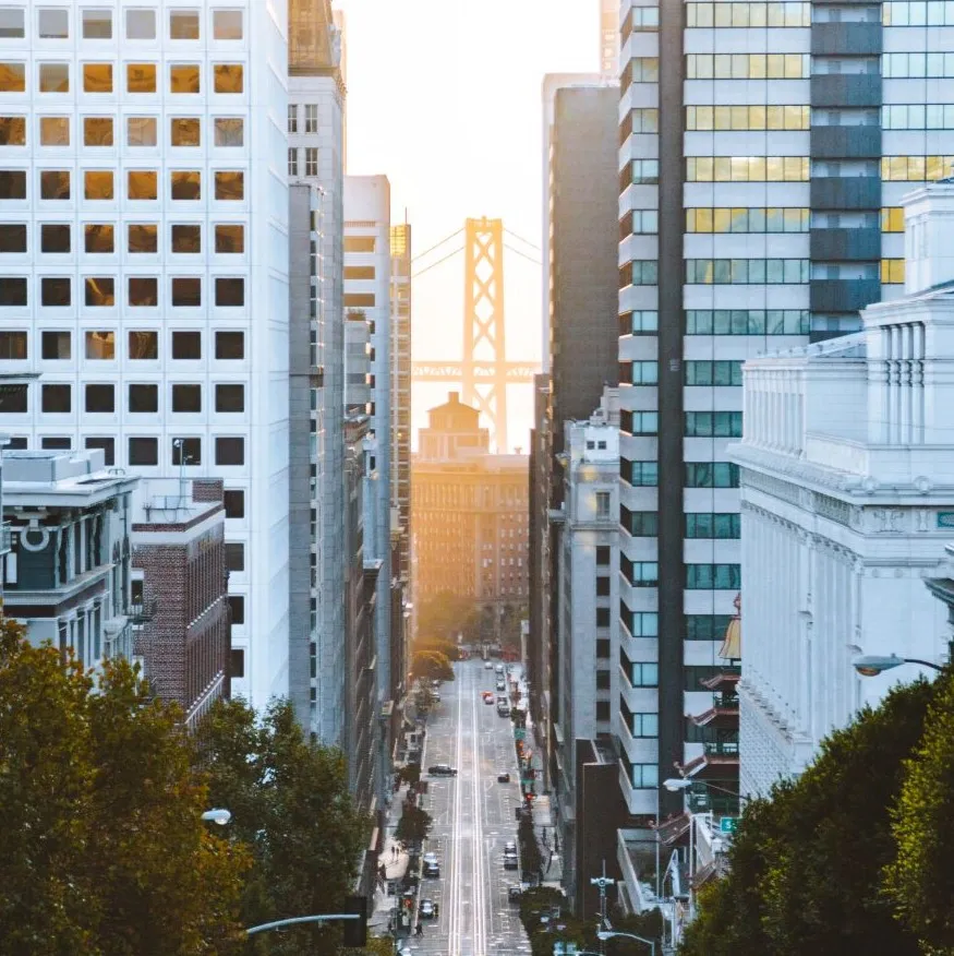View down a city street lined with tall buildings on both sides, leading towards a large suspension bridge in the background, bathed in warm sunlight.