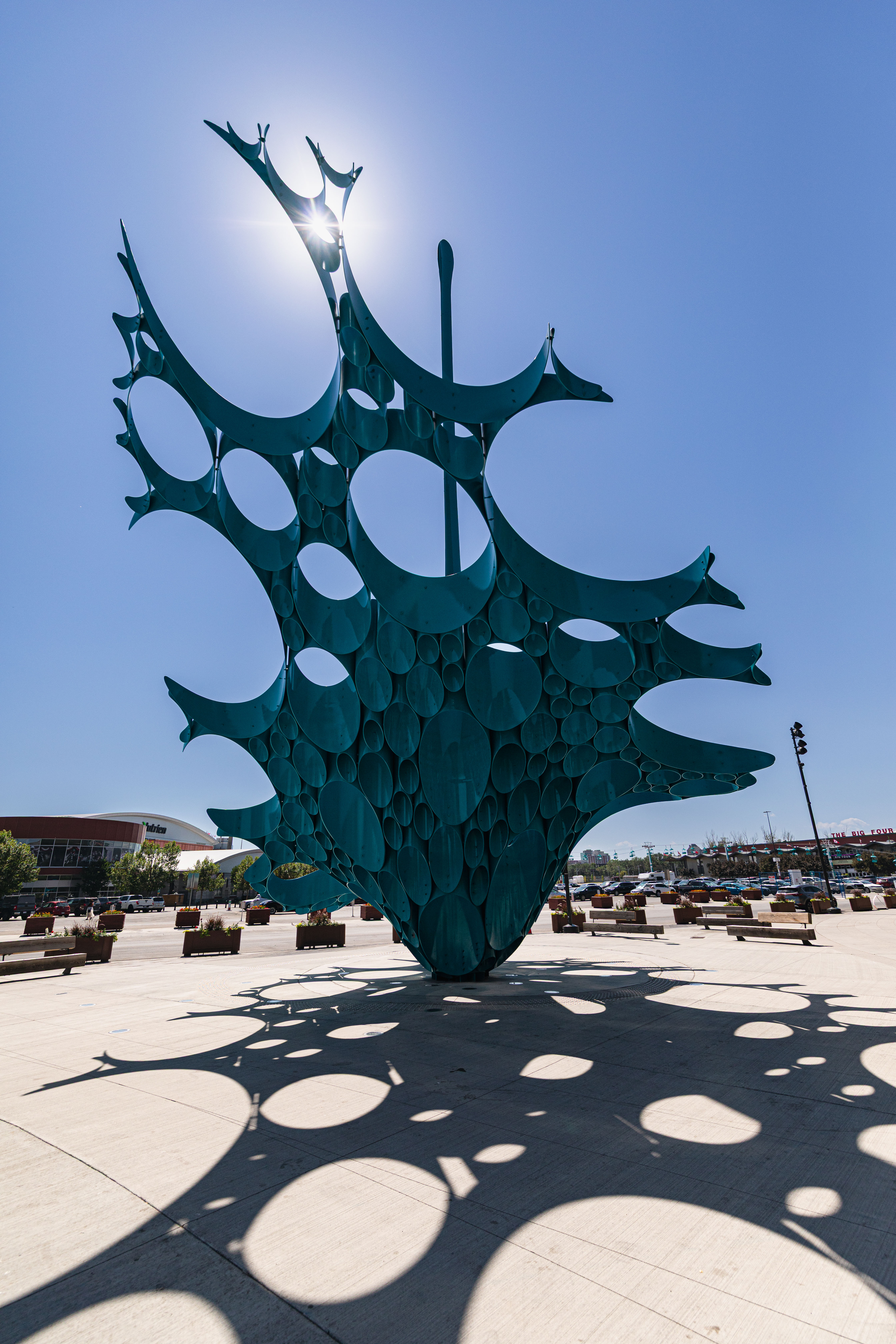 Large outdoor metal sculpture with circular cutouts casting patterned shadows on pavement under bright sun.