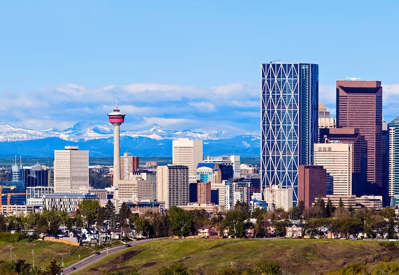 City skyline with tall buildings and a tower, backed by snow-capped mountains under blue sky.