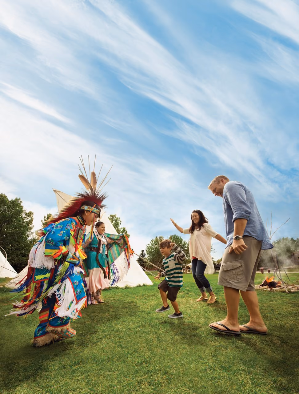 A Native American dancer in colorful traditional regalia dances with a woman, a young boy, and a man on grass near teepees and a campfire under a cloudy blue sky.