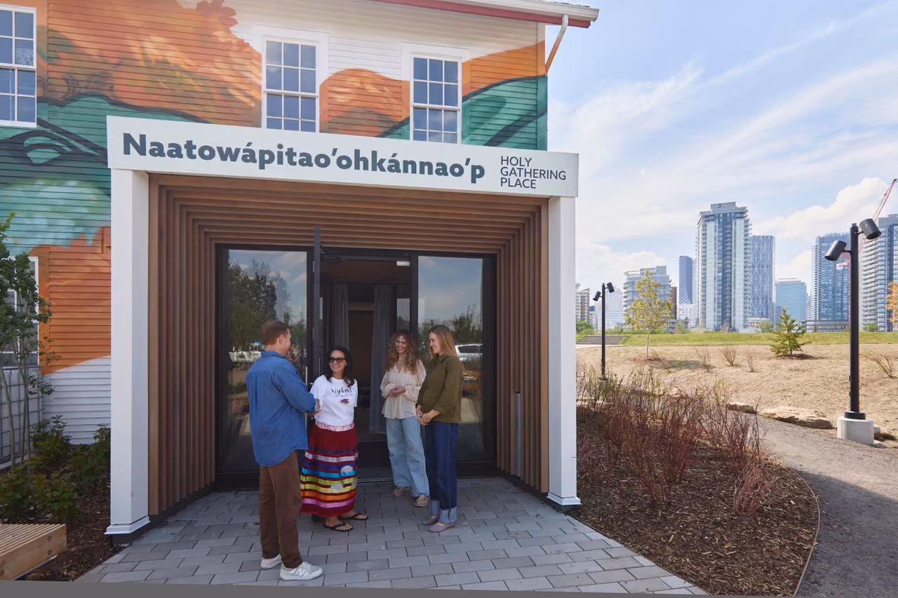Four people standing and talking in front of an entrance with sign reading 'Naatowápitao’ohkánnao’p Holy Gathering Place' on a building with colorful mural and city skyline in background.