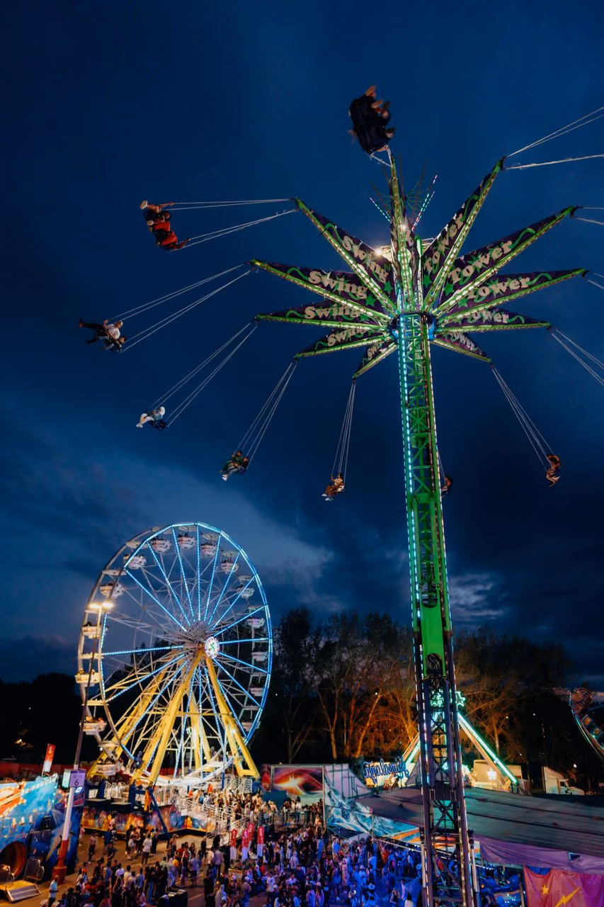 Nighttime carnival scene with people on a brightly lit swing ride tower and a large Ferris wheel glowing in the background over a crowd of visitors.