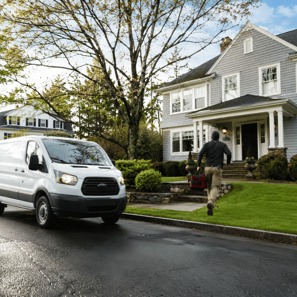 Man carrying tool bag walking from a white van towards a residential grey house with a porch.