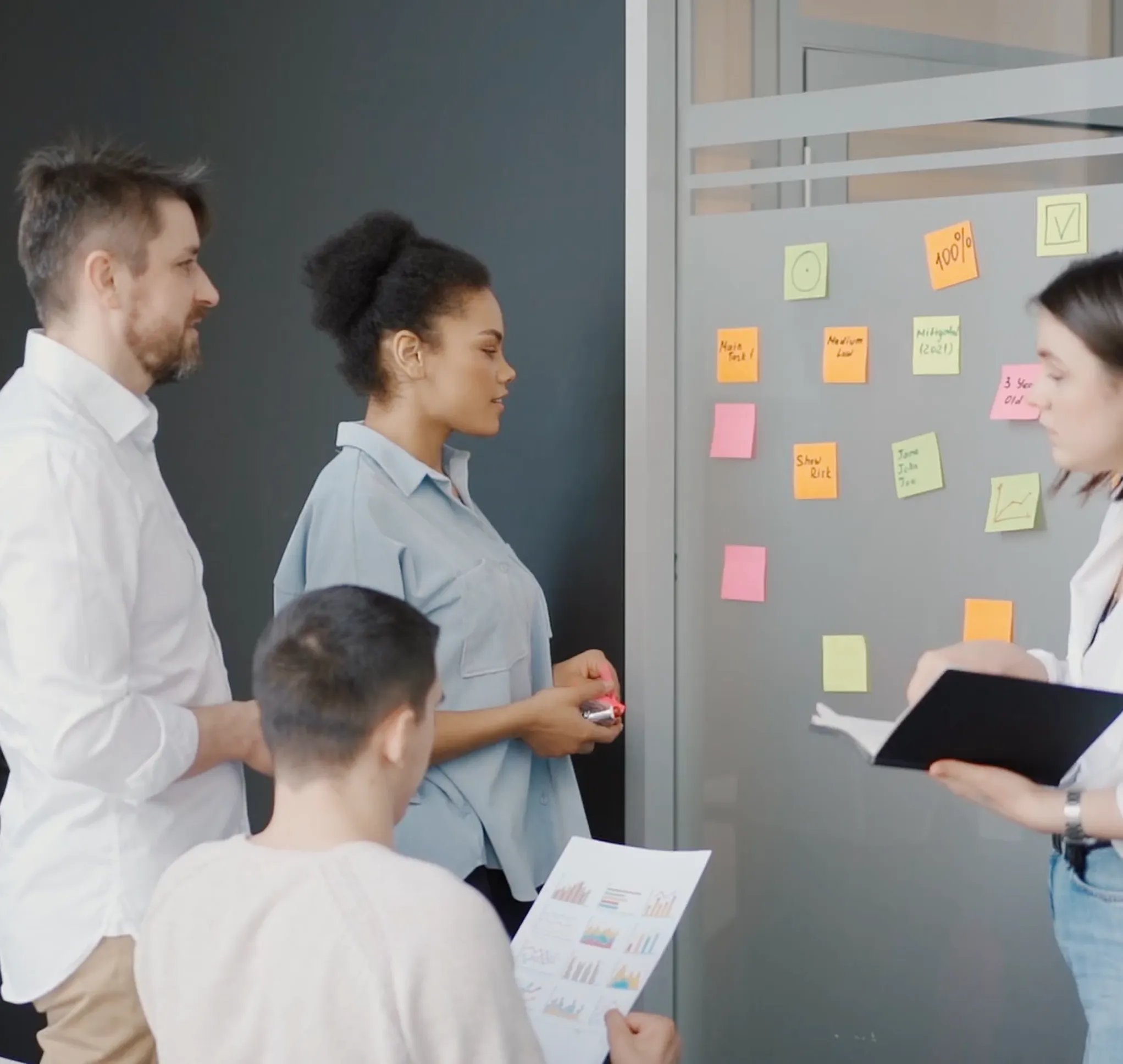Quatre collègues organisent un projet autour de notes affichées sur une paroi vitrée.