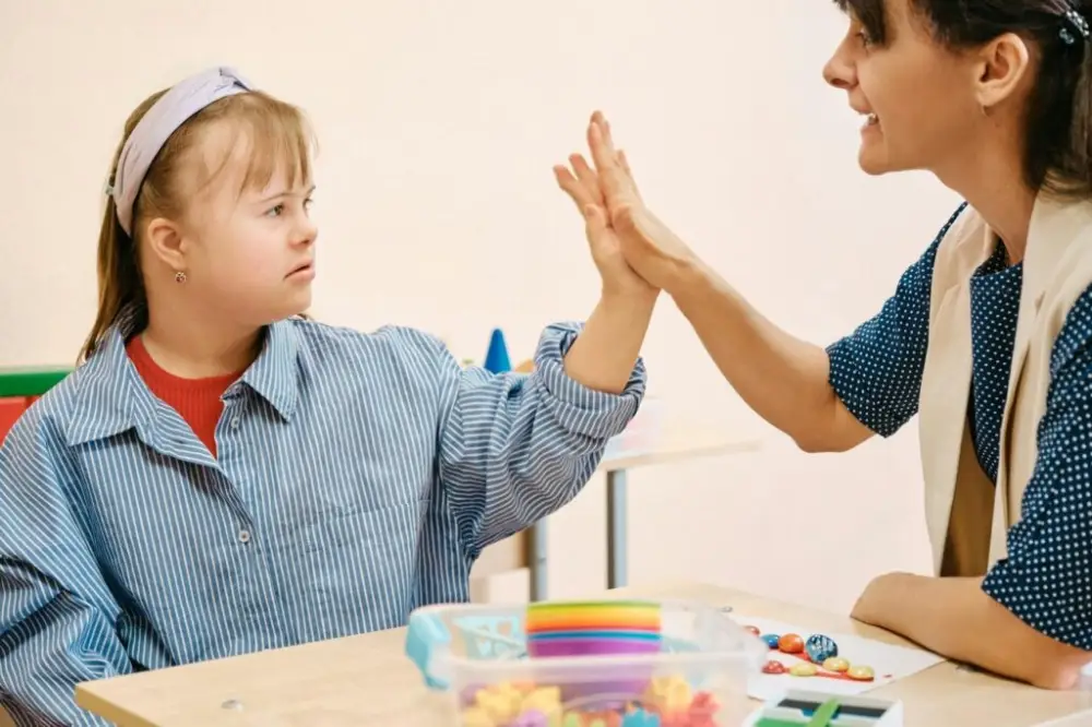 Niña con síndrome de Down chocando las manos con una terapeuta en una mesa con juguetes.