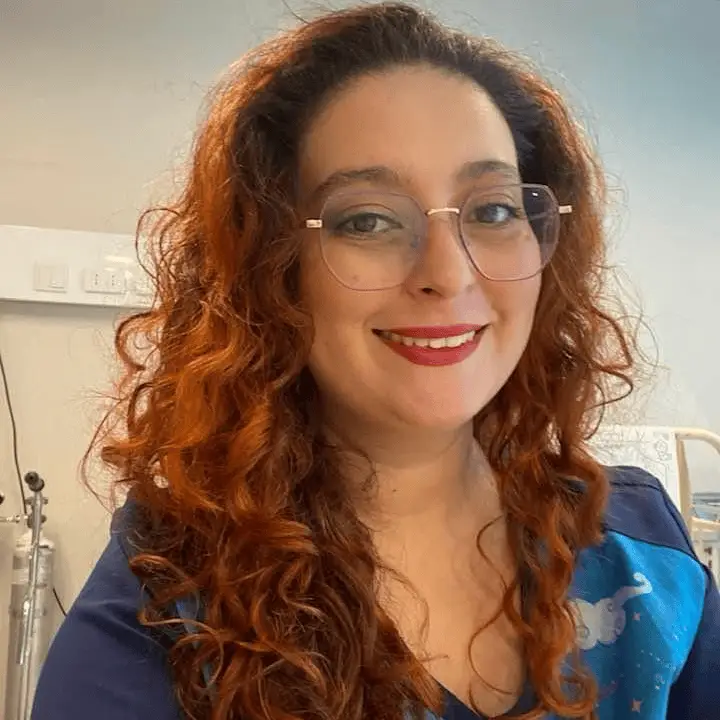 Retrato de Fernanda. Mujer con cabello rizado rojo y gafas grandes, sonriendo en un entorno interior.