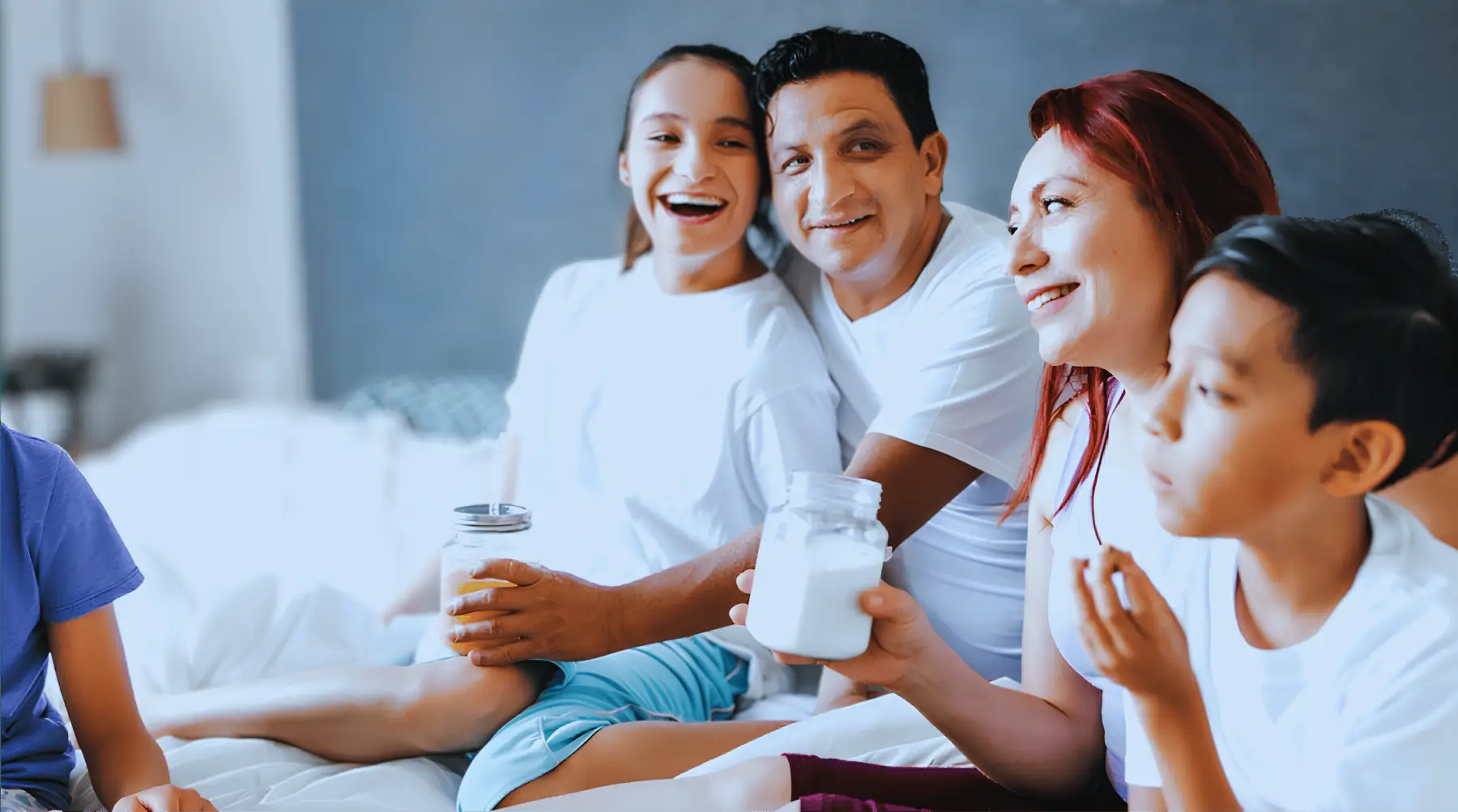 Familia sonriente sentada junta en cama, madre y padre sostienen frascos, niño come galleta.