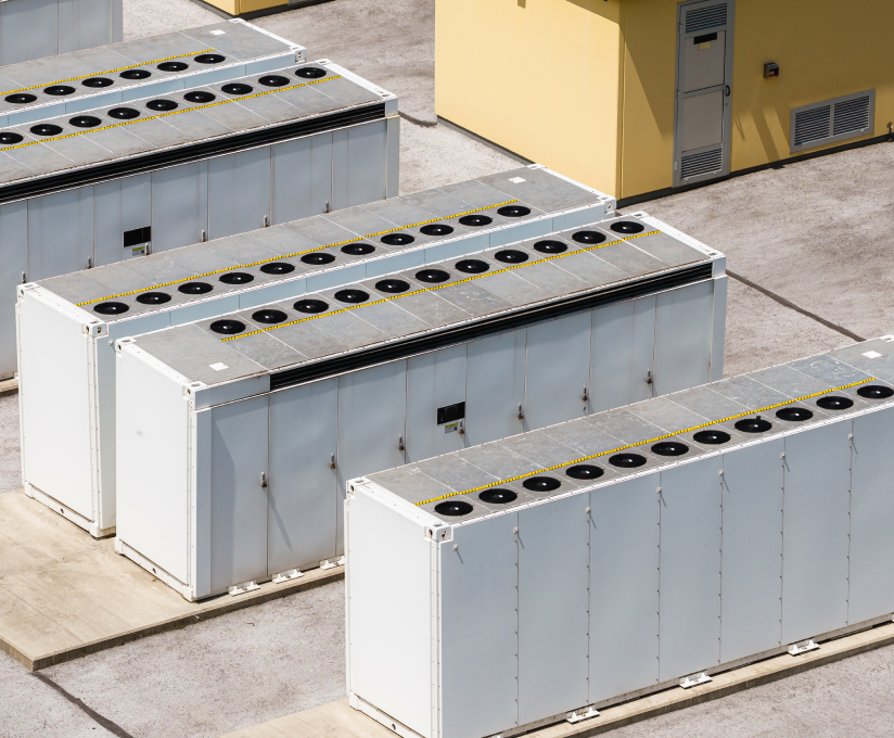Several large white and gray industrial battery energy storage units outdoors near a yellow building.