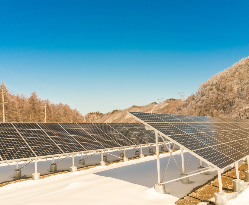 Rows of solar panels installed on snow-covered ground amid hilly, forested landscape under clear blue sky.