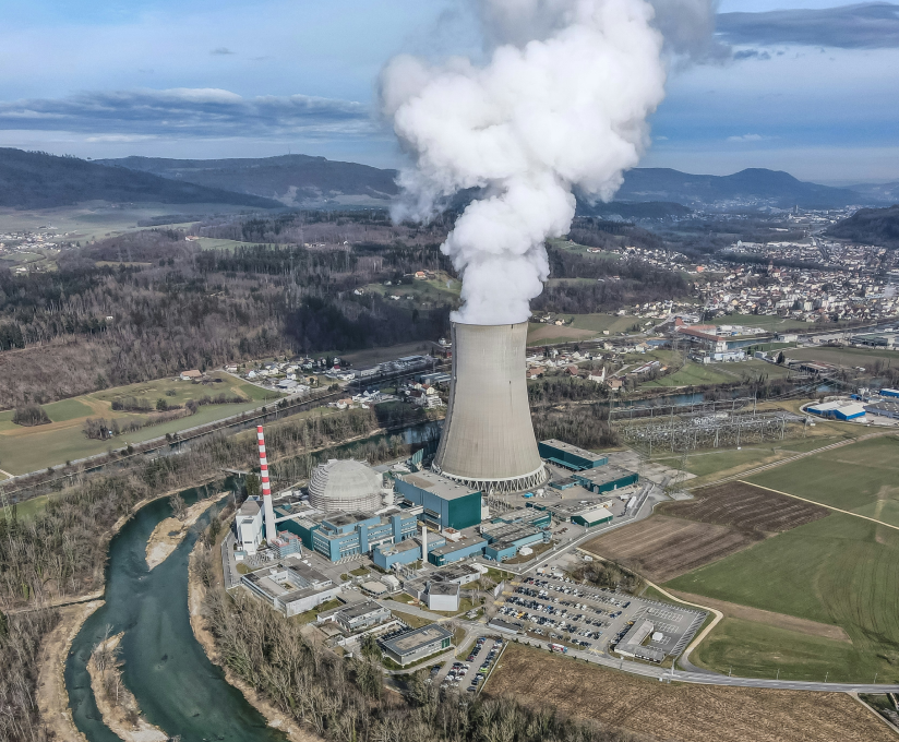Aerial view of a nuclear power plant with a cooling tower emitting white steam near a river and fields.
