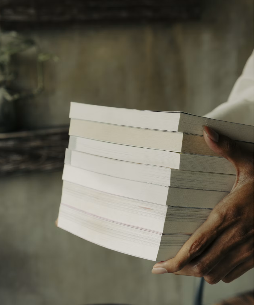 Person holding a stack of thick paperback books with neutral gray background.
