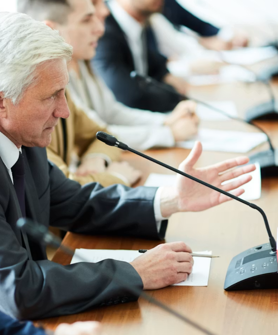 Older man in suit speaking at conference table with others, gesturing with hand near microphone.