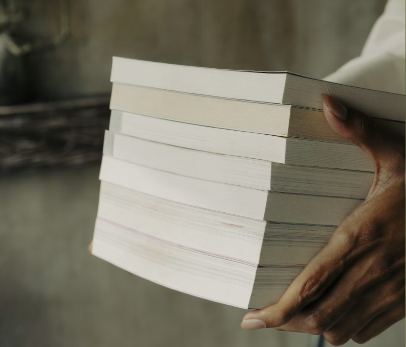 Hand holding a thick stack of white paperback books against a blurry background.