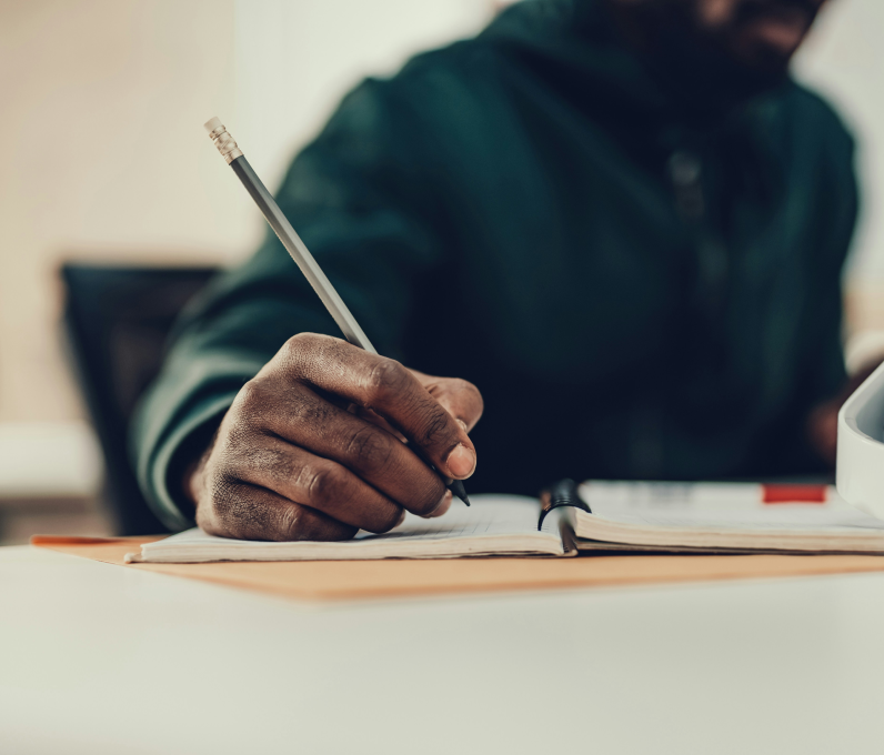 Person writing with a pencil in an open notebook on a desk.