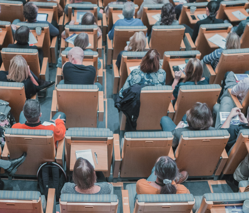 Audience seated in rows of wooden chairs, some using laptops and taking notes during an event.