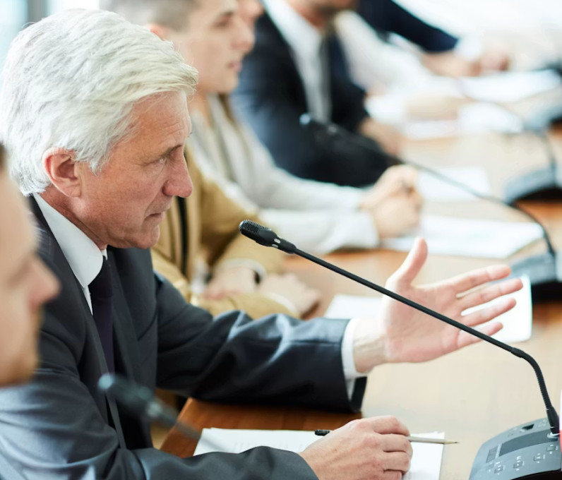 Mature man in suit speaking into microphone during a meeting with colleagues at conference table.