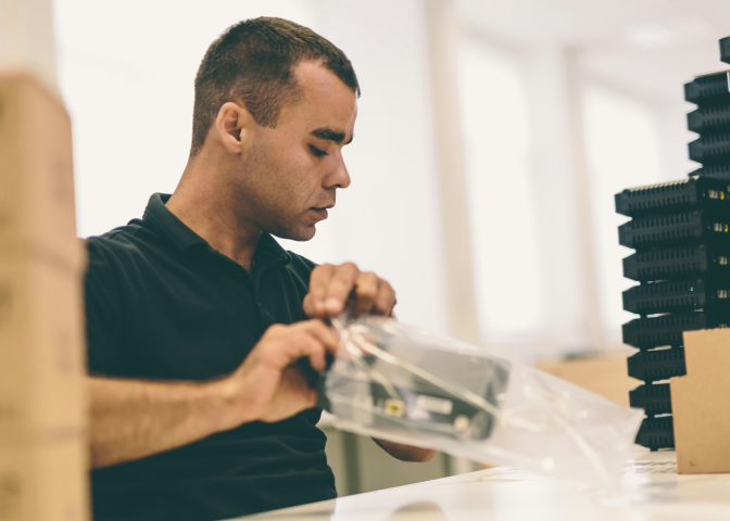 Man in black shirt packaging electronic device in clear plastic bag at a desk in bright room.