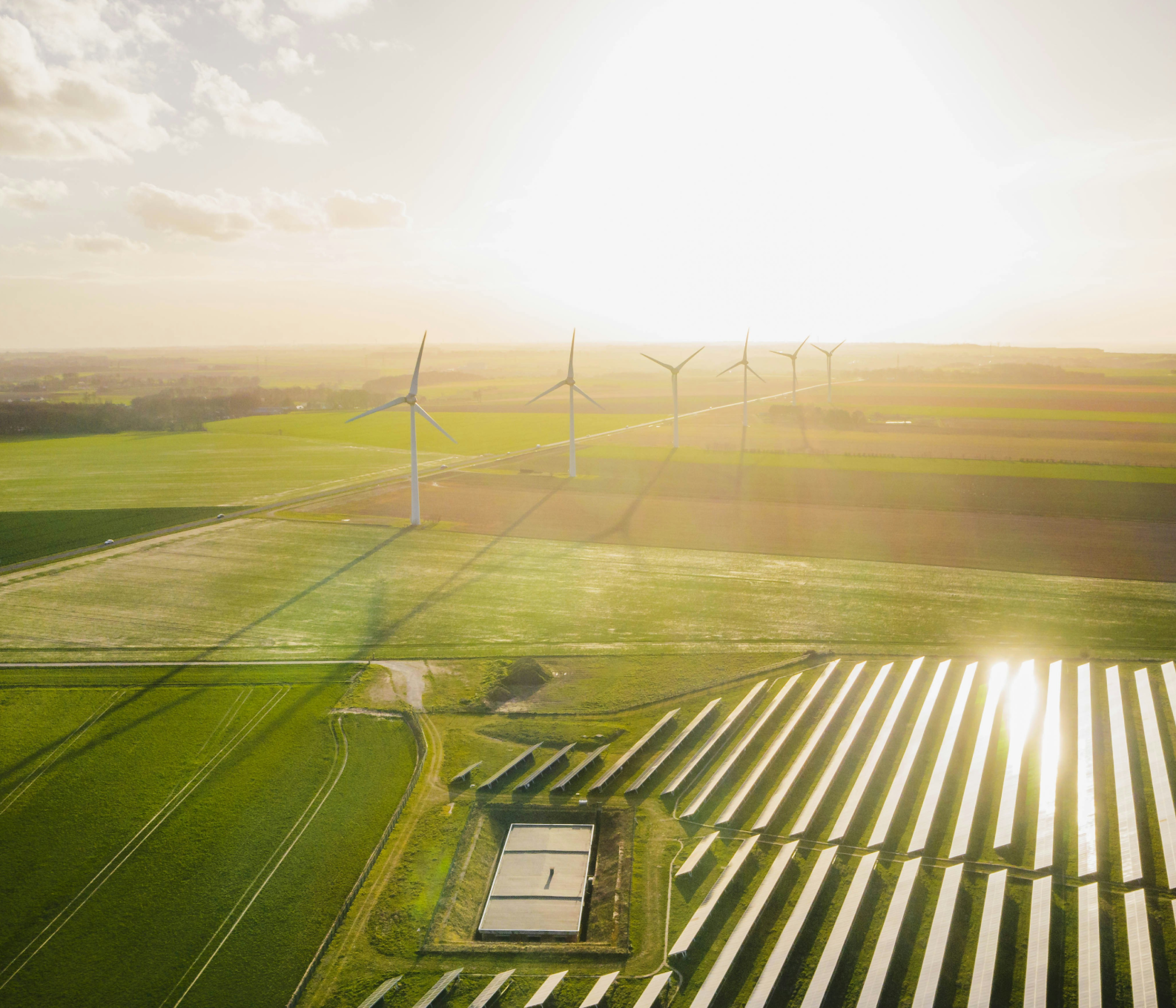 Aerial view of solar panels and wind turbines in green fields under a bright sky at sunset.