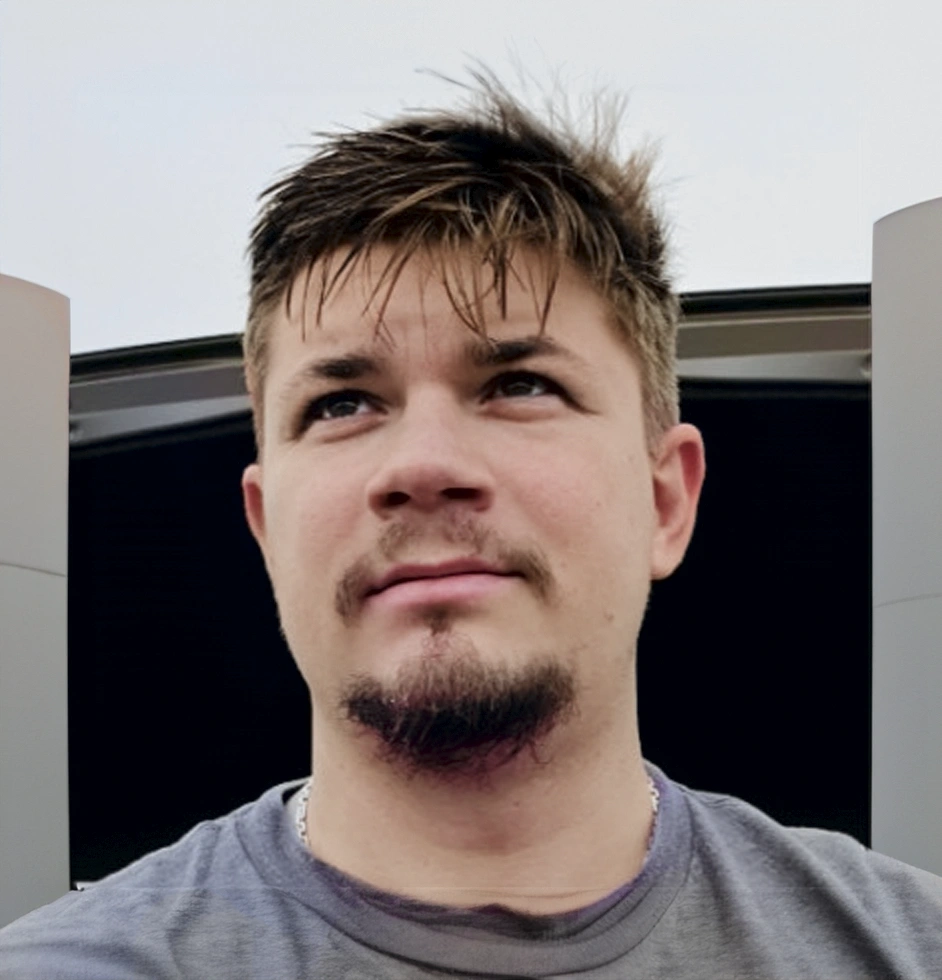 Young man with short brown hair and goatee looking slightly upward outdoors.