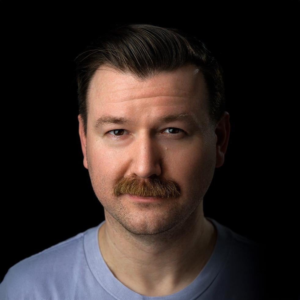 Portrait of a man with brown hair, mustache, and a light blue shirt against a black background.