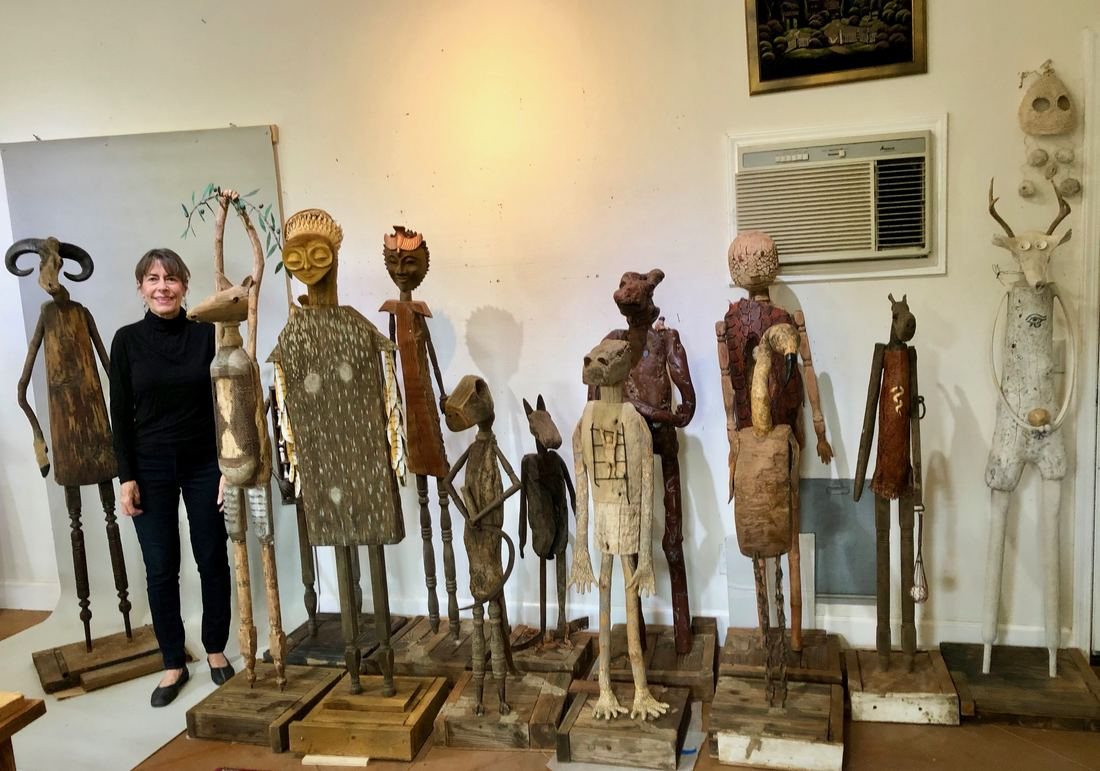 Ann Savageau standing in warm light beside the whole group of driftwood Guardians figures in her studio