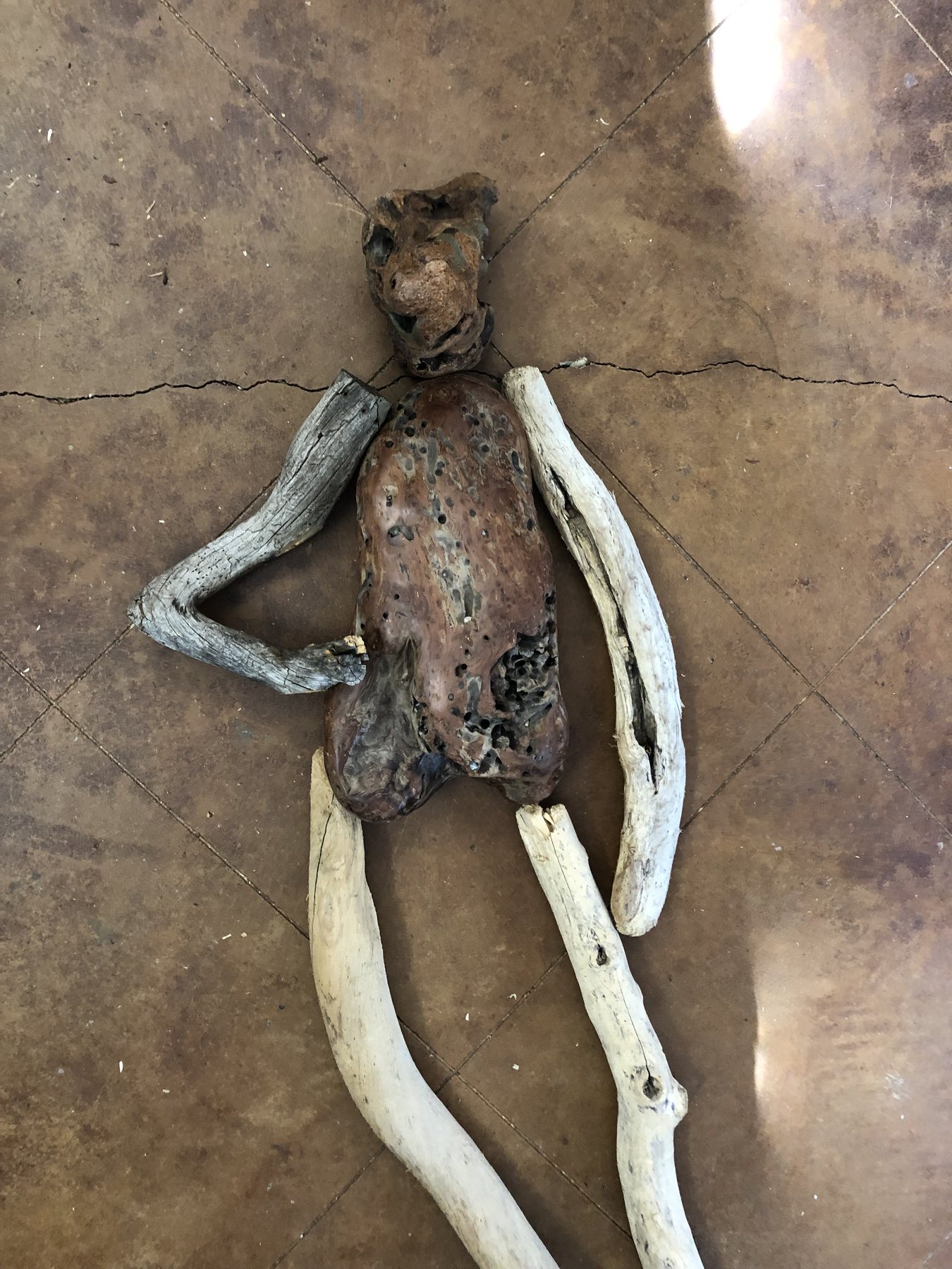 An unfinished figure lying flat on a tiled studio floor, its limbs made of driftwood