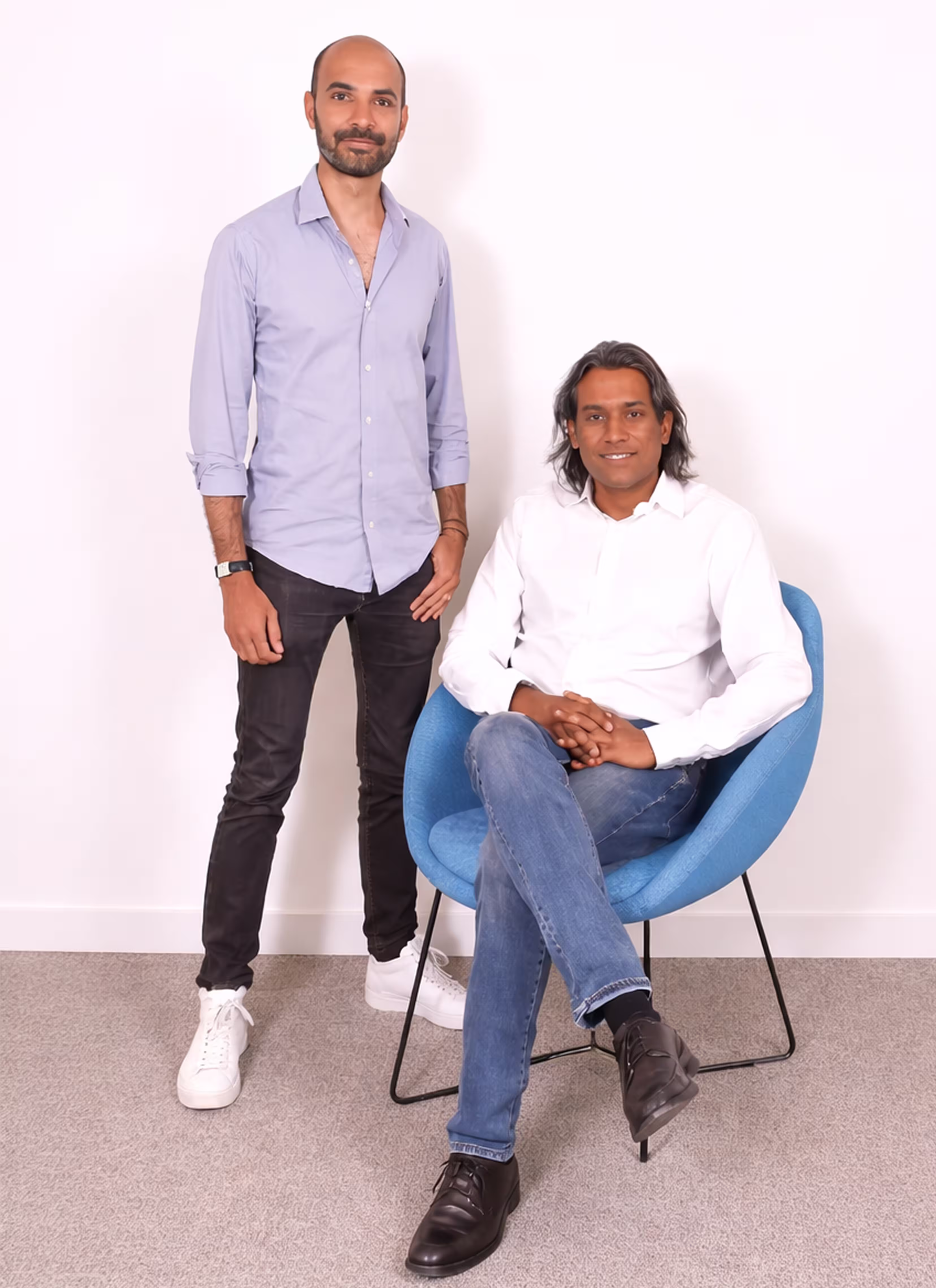 Two men posing against a white wall; one stands wearing a light blue shirt, black pants, and white sneakers, the other sits in a blue chair wearing a white shirt, blue jeans, and black shoes.