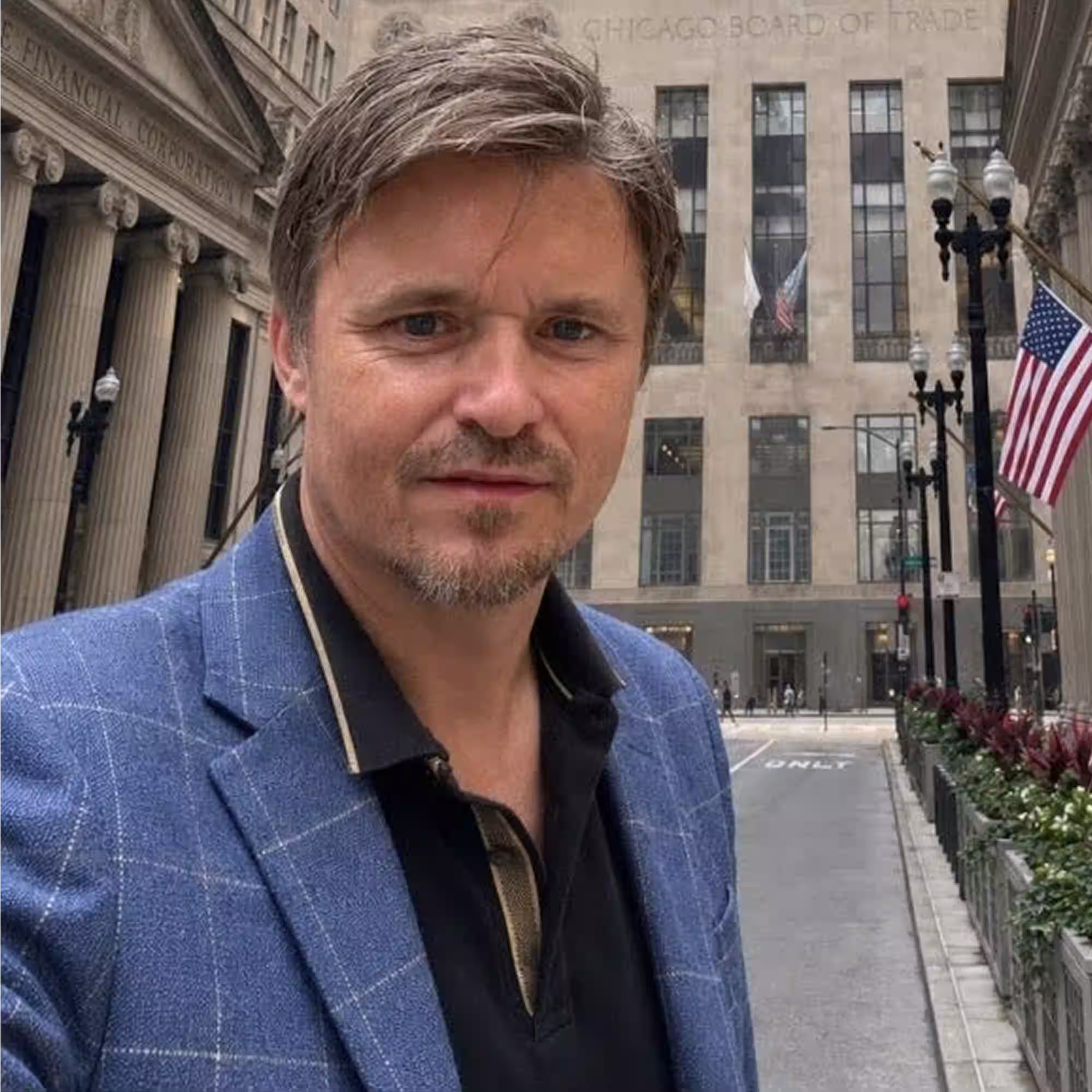 Man with short light brown hair and a trimmed beard wearing a blue checkered blazer and black shirt standing outdoors with the Chicago Board of Trade building and American flags in the background.