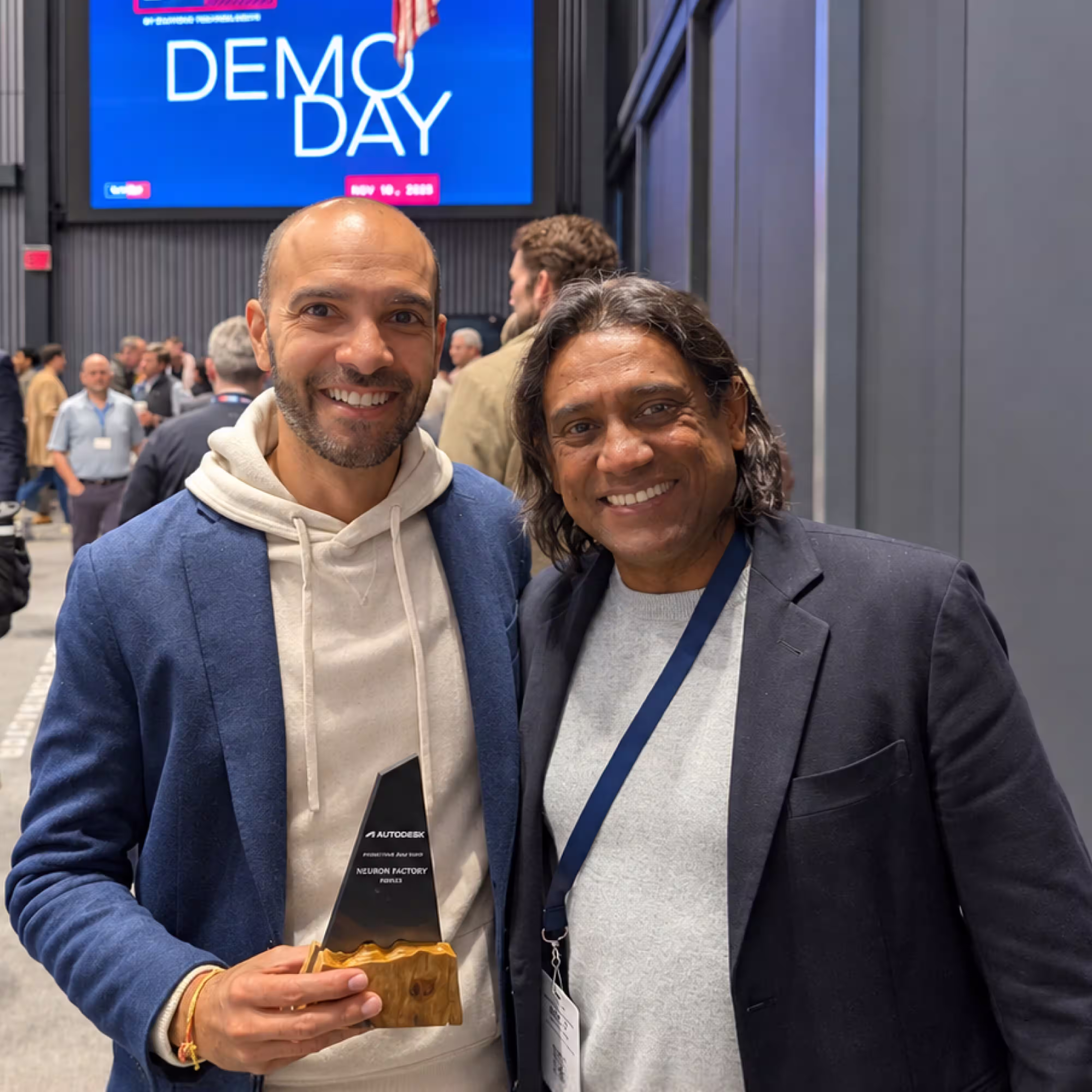 Two smiling men posing indoors at a business event with a blue screen in the background displaying 'DEMO DAY.' One man on the left holds an Autodesk Innovation Award trophy for Neuron Factory.