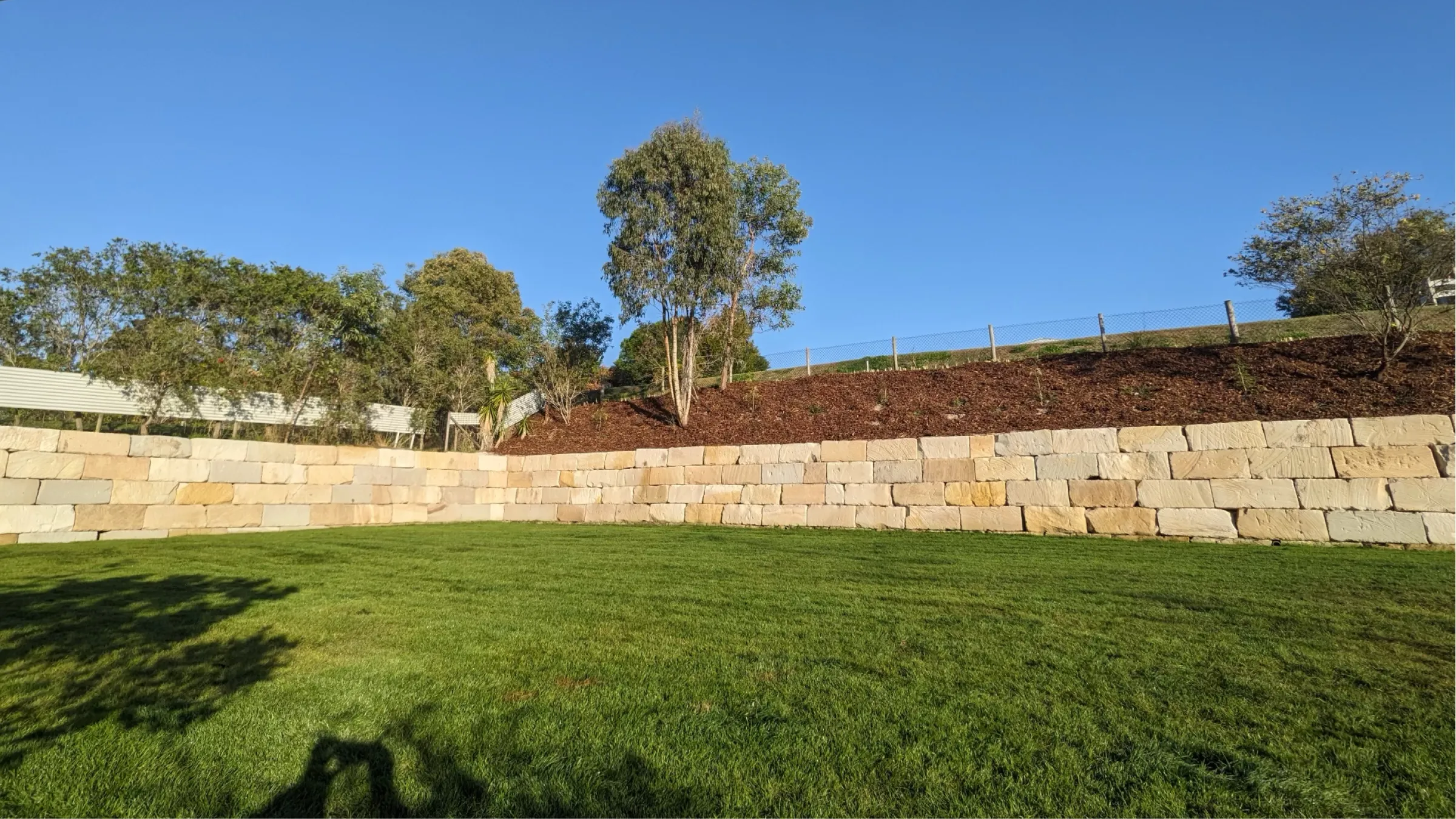 A neatly trimmed green lawn bordered by a stone retaining wall with trees and blue sky above.