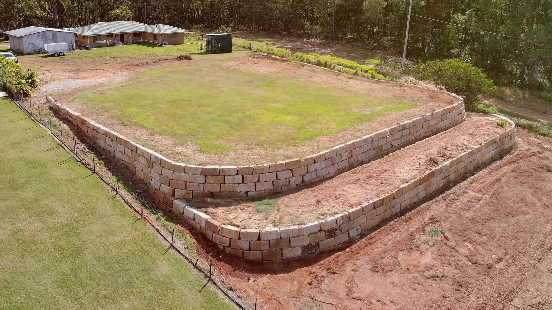 Two-tiered stone retaining wall with grass on top and a dirt area below beside a house in a wooded area.