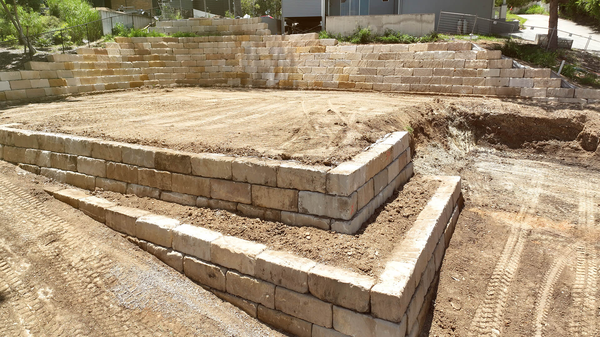 Multi-level retaining wall made of large beige stone blocks with dirt areas and tire tracks around.