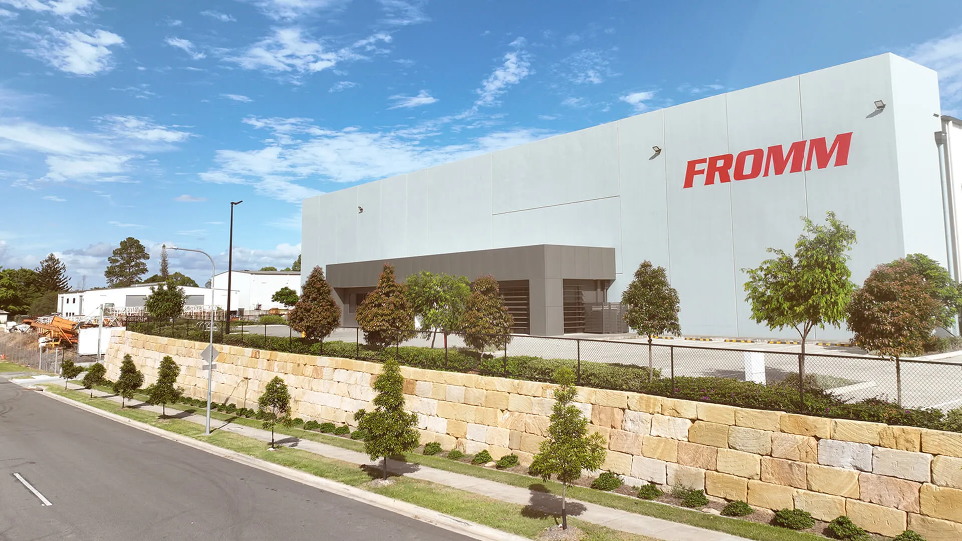 Large FROMM building with surrounding trees, stone wall, and clear blue sky.