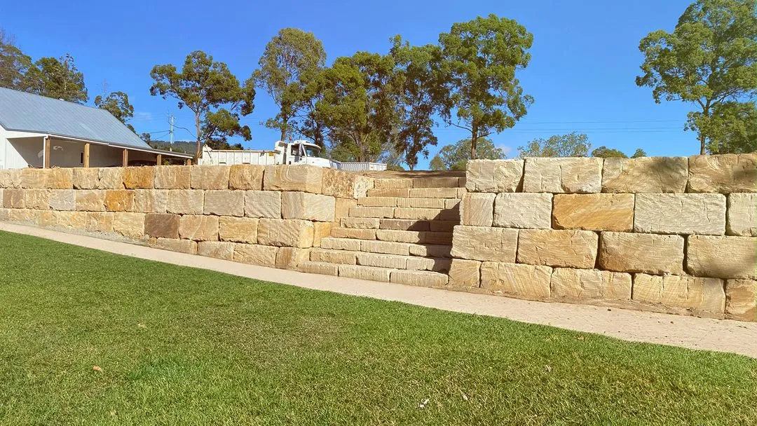 Stone retaining wall with built-in stone steps, grassy area in front, trees and truck behind under blue sky.
