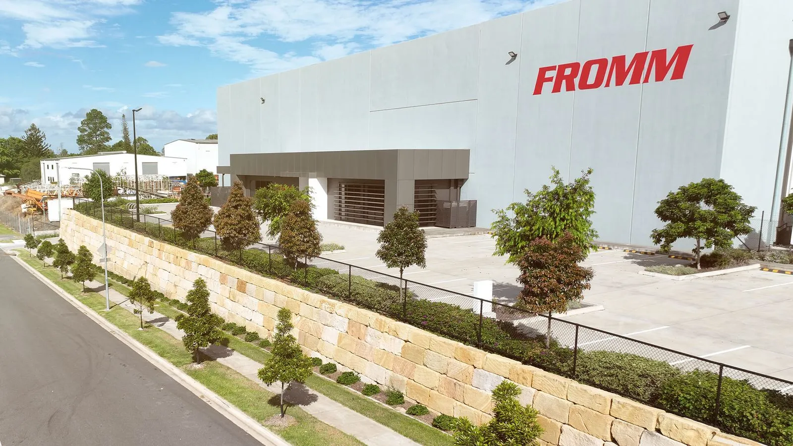 Modern building with FROMM logo, landscaped with trees, stone wall, and an empty parking lot.