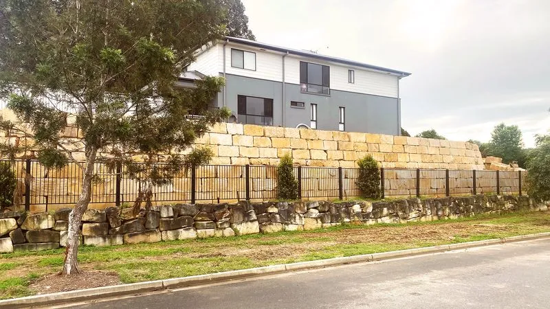 Modern two-story house with grey and white exterior behind layered stone retaining walls and a metal fence.
