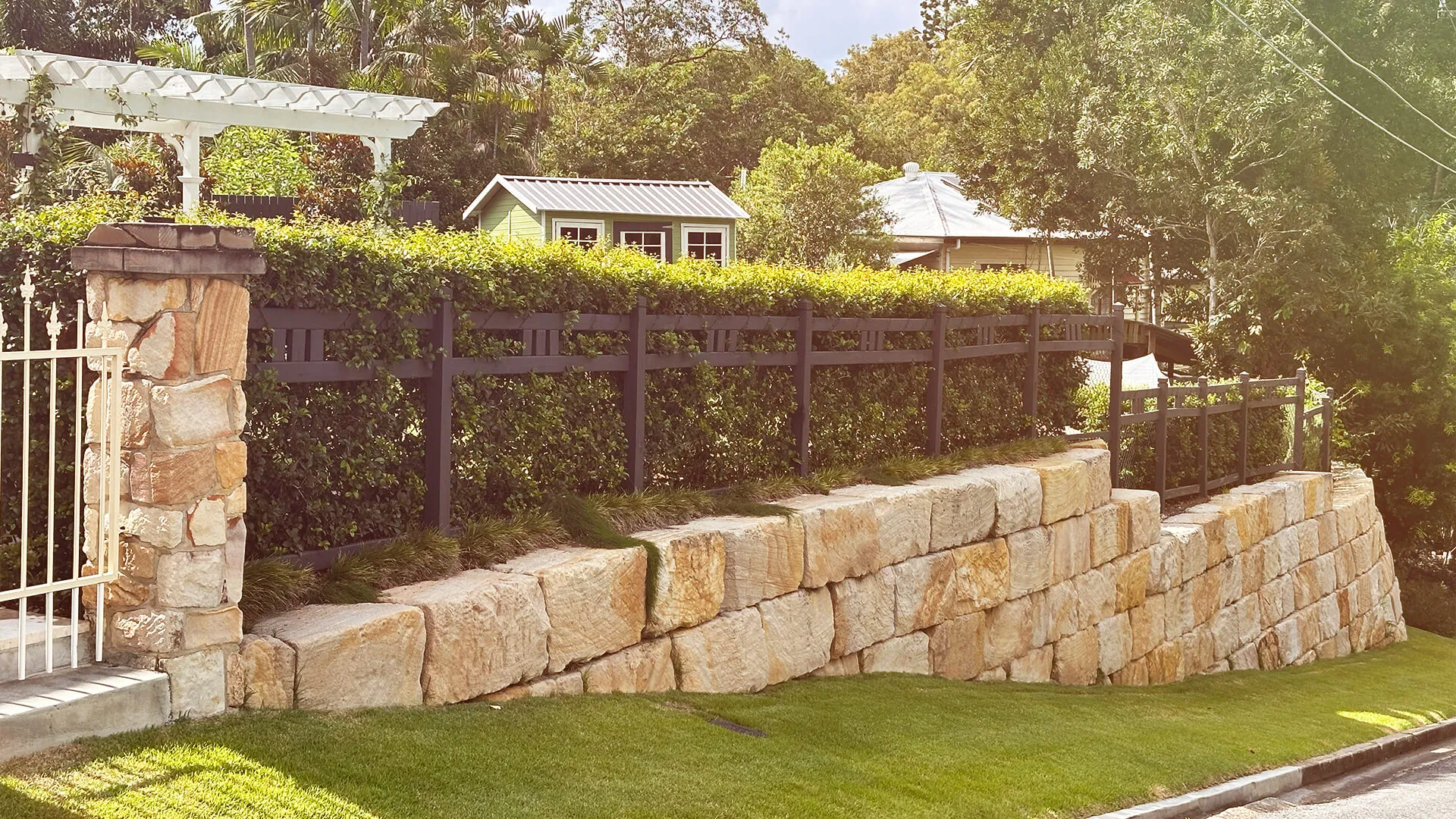 Stone retaining wall with black wooden fence and green hedge behind it in a sunny residential area.