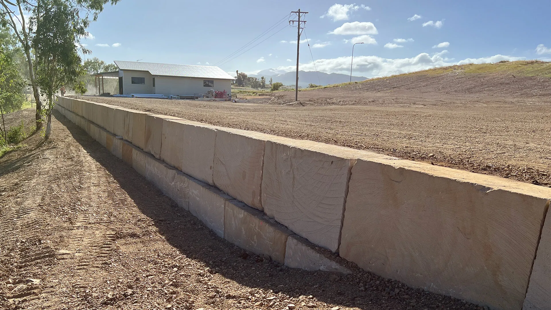 Newly constructed retaining wall made of large stone blocks beside a dirt pathway near a house.