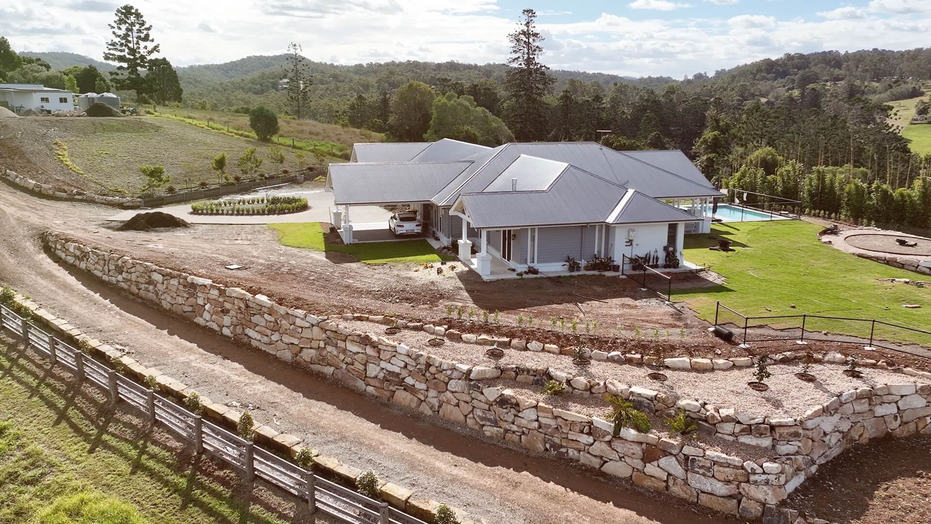 Modern single-story house with gray roofs, stone retaining wall, a driveway, and surrounding greenery.