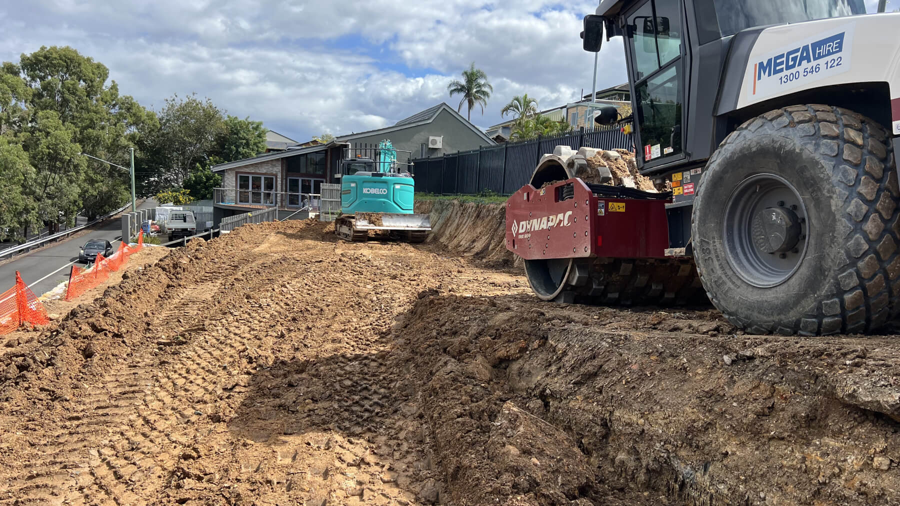 Construction site with dirt being leveled by a Dynapac roller and a Kobelco excavator near houses.